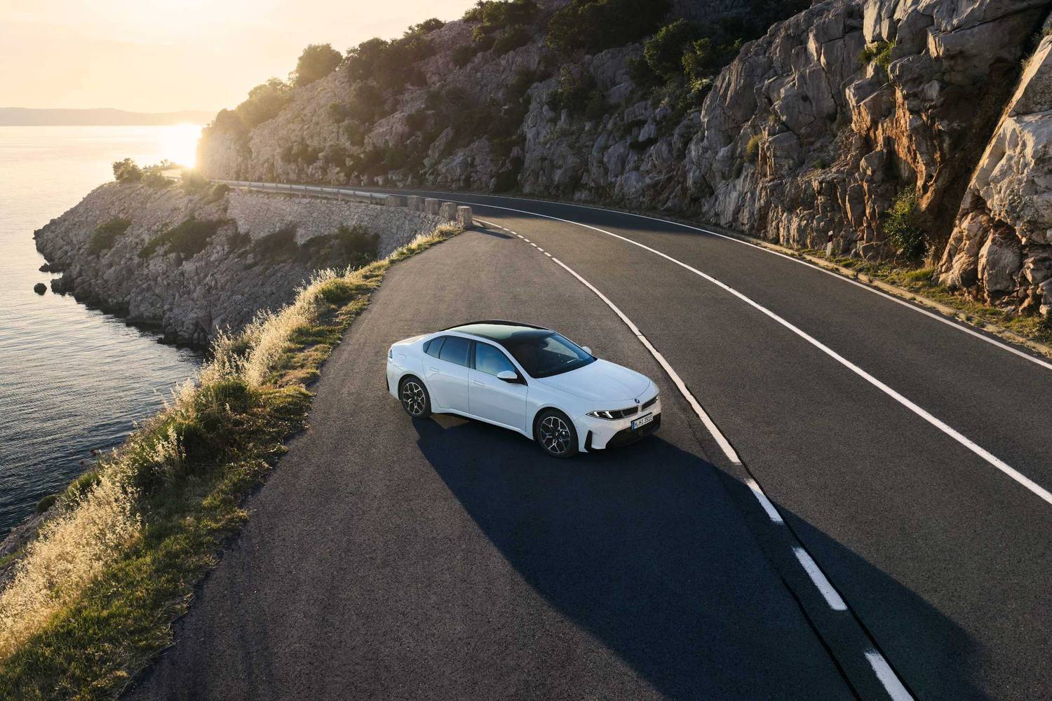 Białe BMW i3 50 xDrive na nadmorskiej drodze o zachodzie słońca